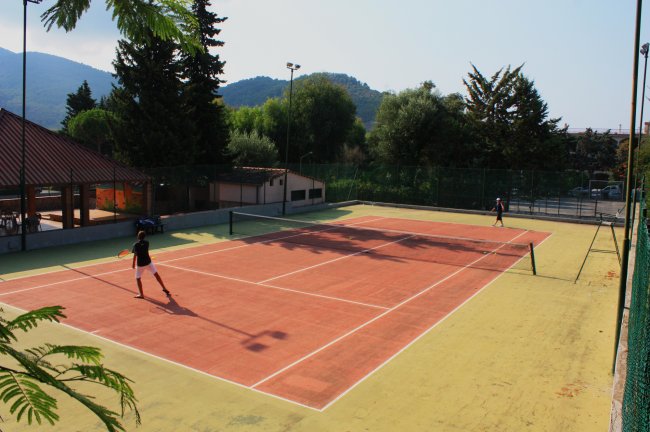 Campo da tennis all'aperto con due persone che giocano, circondato da alberi e colline.