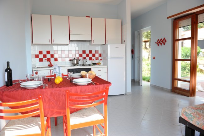 Cucina con tavolo apparecchiato, sedie rosse, e mobili bianchi. Finestra aperta sul giardino.