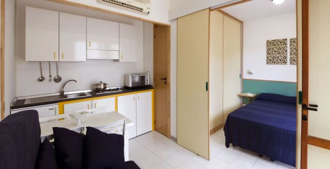 Cucina e area letto con parete divisoria scorrevole; tavolo, sedie e letto singolo sono presenti.