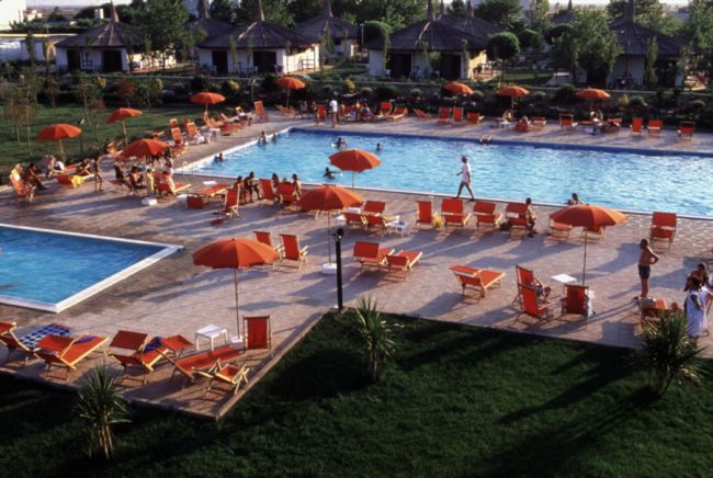 Piscina affollata con sdraio e ombrelloni arancioni, circondata da piccoli edifici e vegetazione.