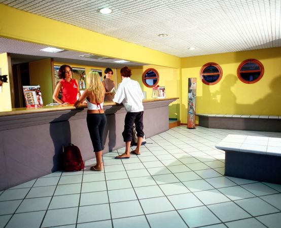 Persone alla reception dietro un bancone in una stanza con pareti gialle e piastrelle bianche.