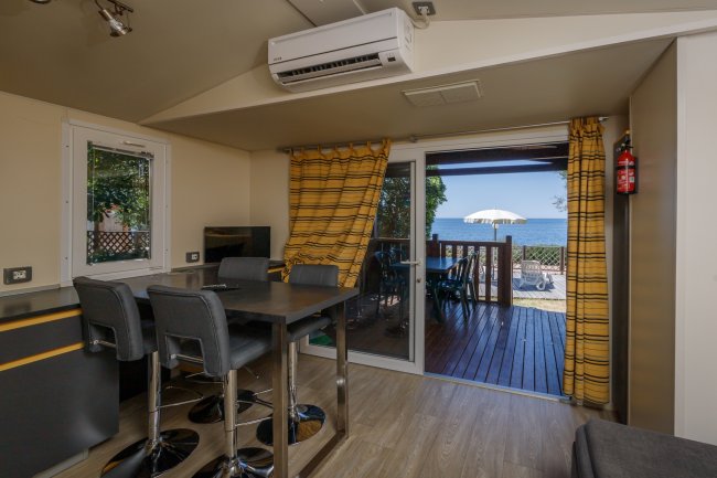Sala con tavolo alto, sedie grigie e vista mare attraverso porta finestra aperta.