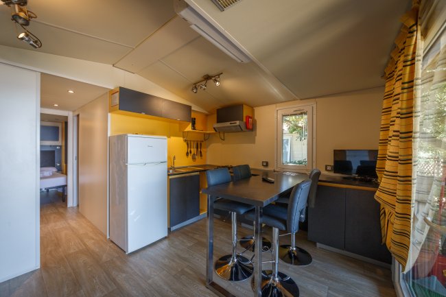 Cucina moderna con tavolo alto, sedie, frigorifero e zona notte sullo sfondo.
