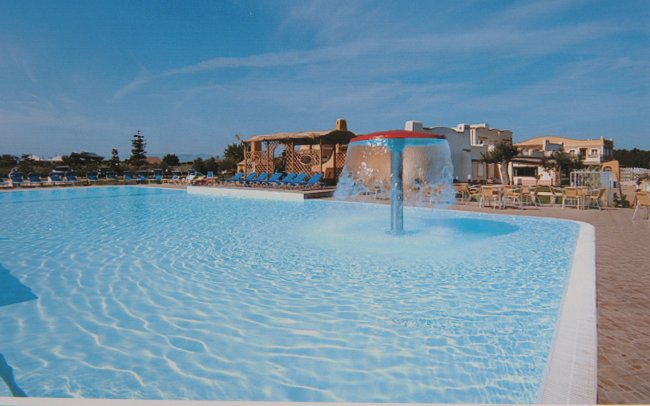 Una piscina con una fontana centrale, circondata da sedie a sdraio e un cielo azzurro.