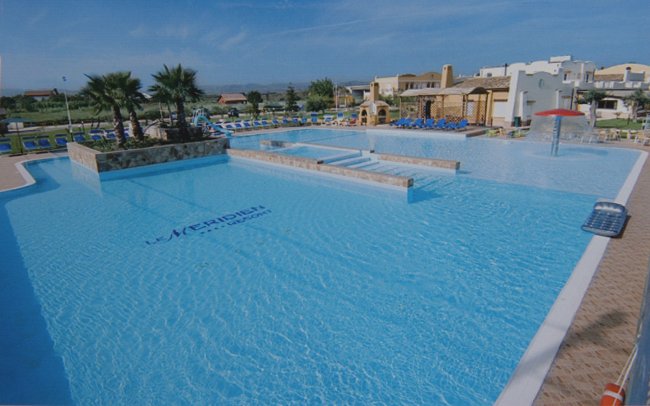 Ampia piscina con fontana e palme, contornata da lettini e edifici bassi.