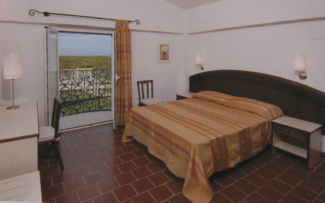 Una camera con letto matrimoniale, pavimento in cotto e balcone aperto su un panorama verde.