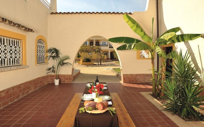 Un tavolo esterno con salumi, pane e vino. Archi, piante tropicali e cortile sullo sfondo.