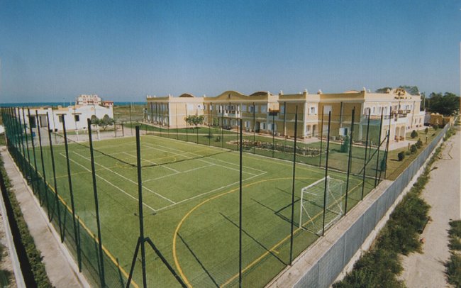 Campo sportivo verde circondato da una recinzione, vicino a edifici gialli e al mare.