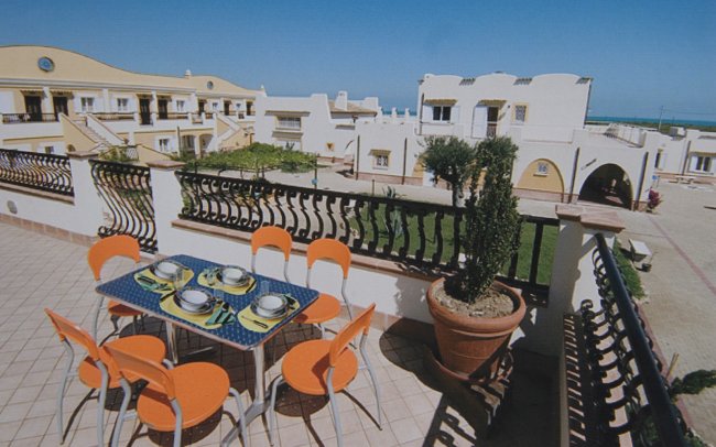 Terrazza con tavolo apparecchiato e sedie arancioni, vista su edifici bianchi.