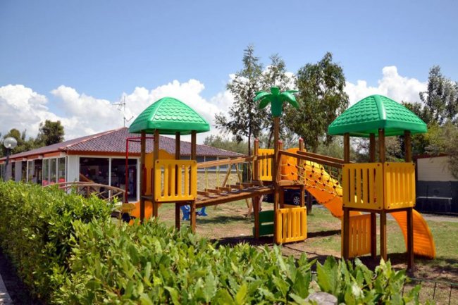 Area giochi all'aperto con strutture gialle e tetti verdi, circondata da vegetazione.