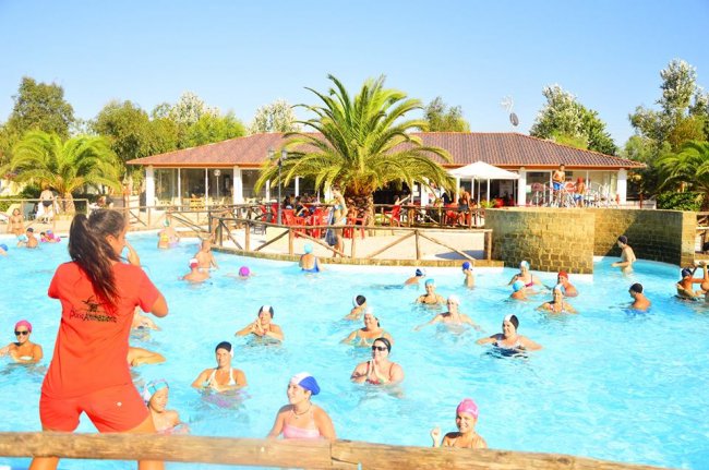 Lezione di acquagym in una piscina all'aperto con partecipanti e istruttrice in divisa rossa.
