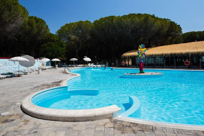 Piscina ampia con un'isola centrale colorata, circondata da ombrelloni bianchi e lettini.
