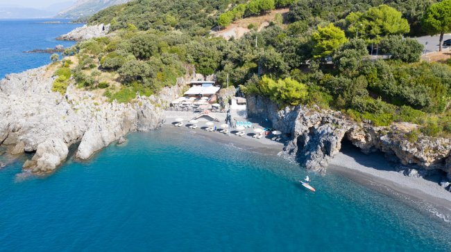 Spiaggia con mare blu e piccola baia rocciosa, ombrelloni e un edificio.