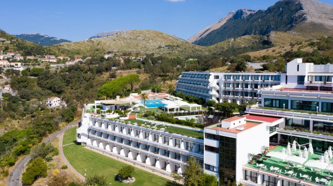 Grande hotel con terrazze, piscina e vista su montagne e colline verdi.