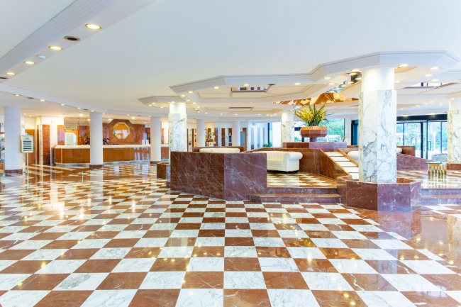 Una hall di hotel con pavimento a scacchi marrone e bianco, colonne di marmo e soffitto luminoso.