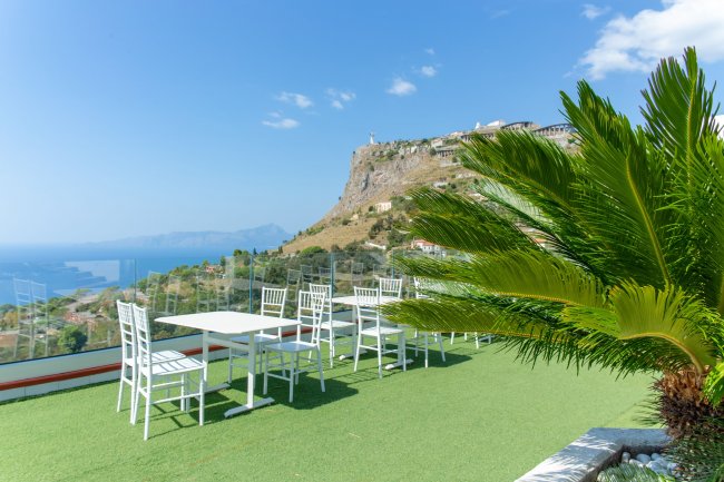 Terrazza con tavolo e sedie bianche, vista su una montagna e mare.