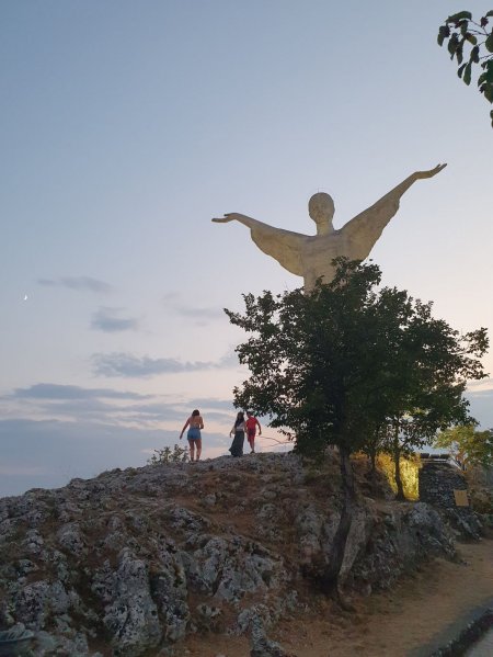 Statua con braccia aperte su collina al tramonto, persone camminano su sentiero roccioso.