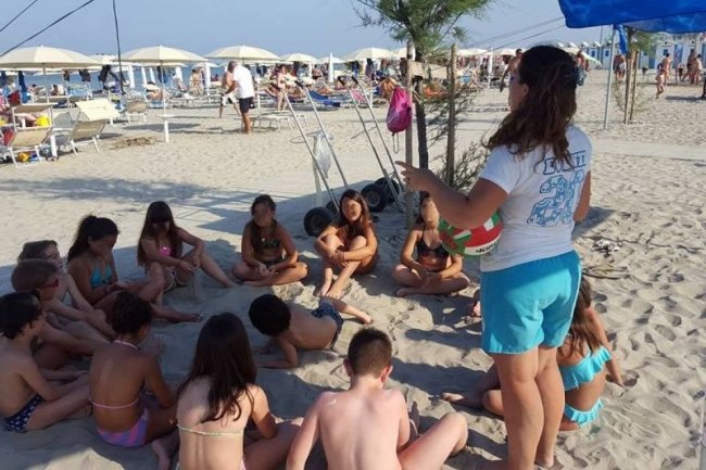 Un gruppo di bambini ascolta una donna sulla spiaggia, circondati da ombrelloni e sedie a sdraio.