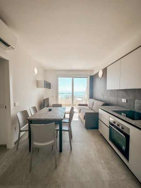 Interno moderno con cucina, tavolo, sedie e vista sul mare da una porta-finestra.