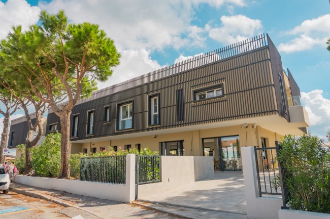 Edificio moderno con rivestimento scuro, circondato da alberi e un cancello.