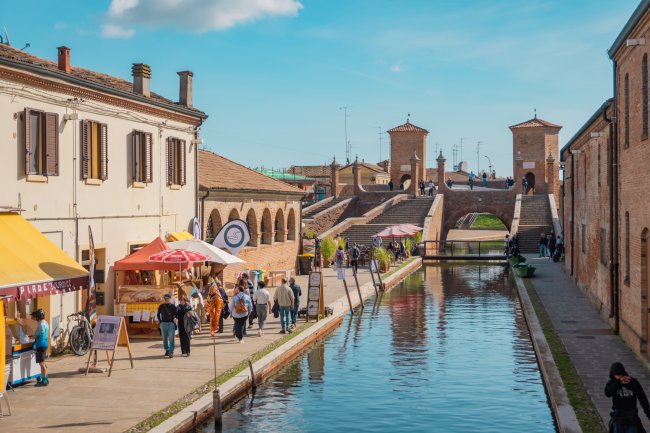 Canale con passerella, ponti in pietra e stand con persone nella città di Comacchio.