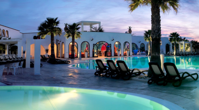 Piscina con arredo elegante e palme al tramonto, circondata da edifici bianchi con vetrate ad arco.
