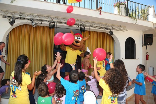 Bambini giocano con palloncini rossi insieme a una mascotte in un'area con tende gialle.