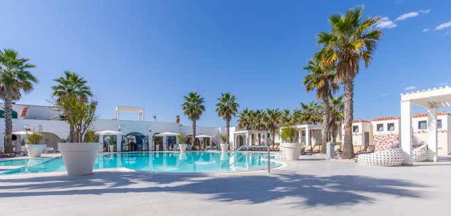 Piscina circondata da palme e lettini, cielo sereno, architettura mediterranea.