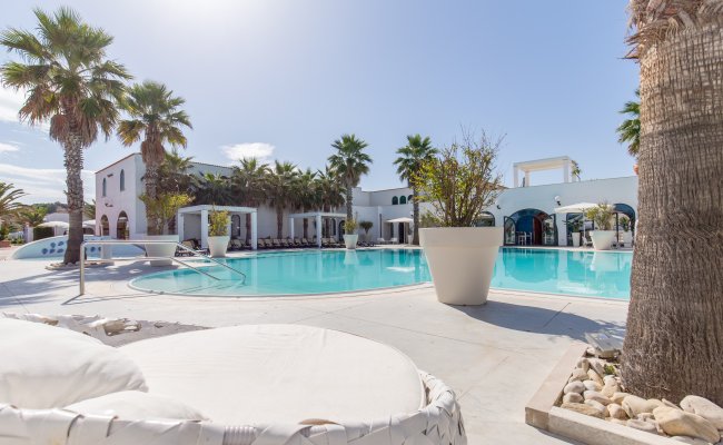Piscina con palme e archi lontani, area relax con cuscini sulla sinistra.