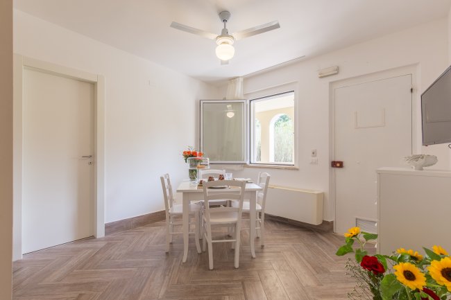 Sala da pranzo con tavolo bianco e fiori. Finestre aperte, pavimento in legno chiaro.