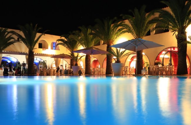 Piscina notturna con palme e ombrelloni illuminati.