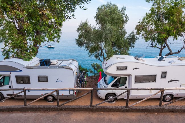 Due camper parcheggiati vicino a un mare calmo, circondati da alberi verdi.