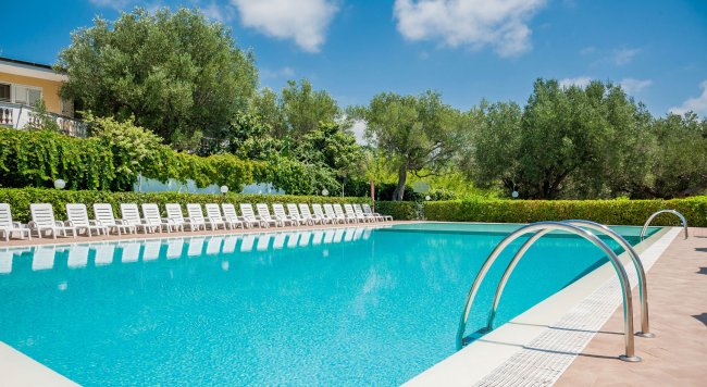 Una piscina all'aperto circondata da sedie a sdraio bianche e alberi verdi sotto un cielo azzurro.