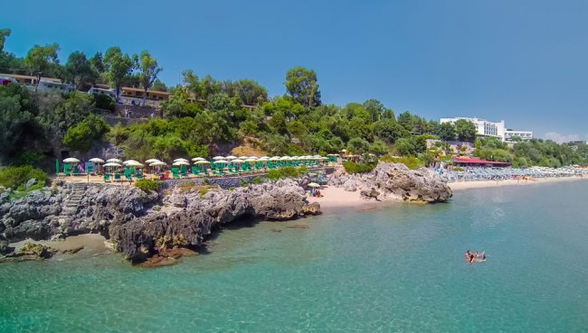 Vista aerea di una spiaggia rocciosa e lettini sotto ombrelloni verdi.