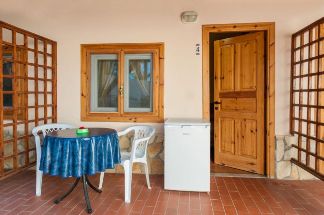 Veranda con tavolo, sedie di plastica, frigo bianco e porta aperta in legno.