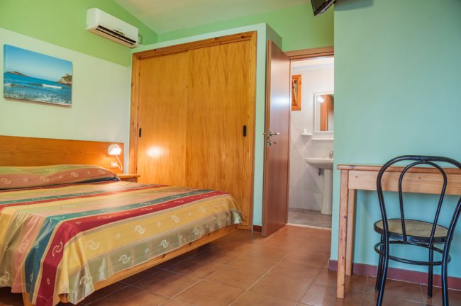 Letto matrimoniale con copriletto a righe colorate, armadio e piccolo bagno in fondo.