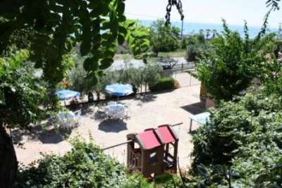Un cortile con tavoli e ombrelloni circondato da vegetazione e vista mare in lontananza.