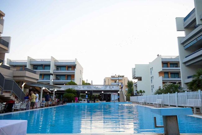 Piscina all'aperto circondata da edifici moderni e persone sedute ai tavolini.