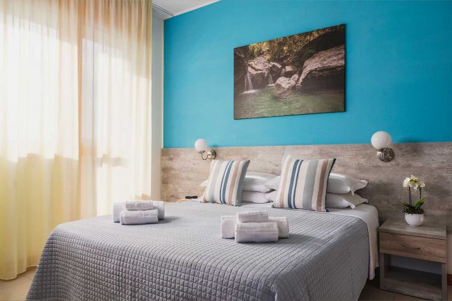 Letto matrimoniale con cuscini a righe, parete blu, quadro sopra il letto e luci a parete.