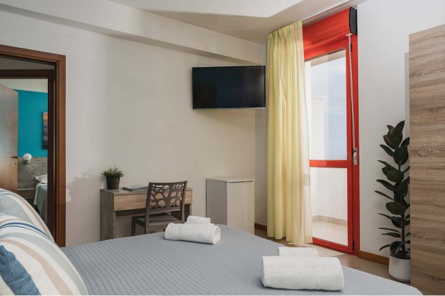 Camera con letto, TV a parete, scrivania, finestra con tenda chiara e porta rossa.