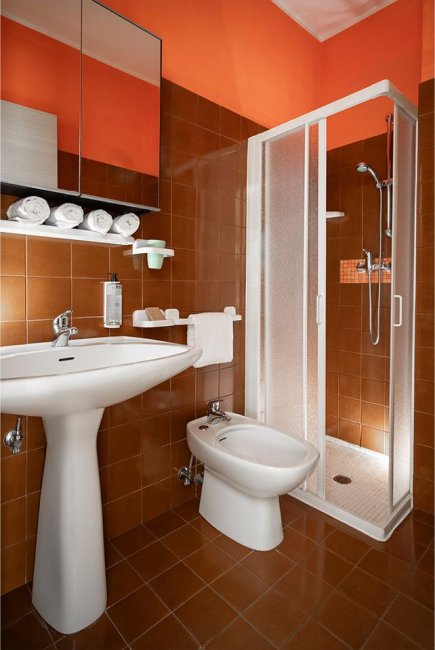 Bagno con piastrelle marroni, lavabo bianco, bidet, doccia chiusa e asciugamani arrotolati.