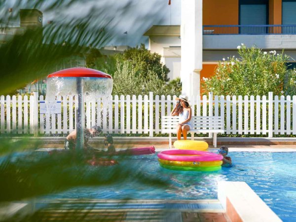Bambini che giocano in una piscina, circondati da recinzioni bianche e piante.