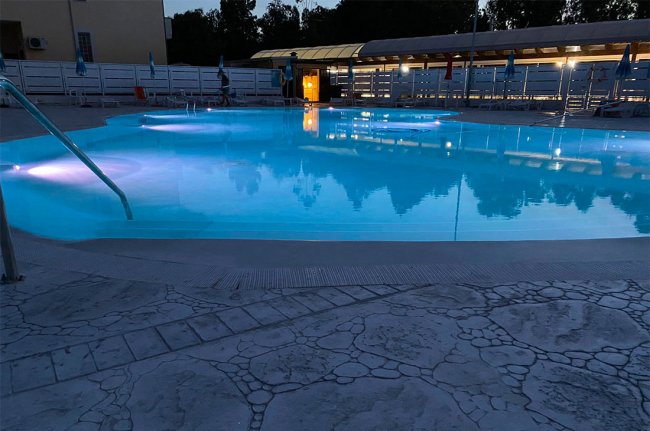 Piscina illuminata di sera con ombrelloni e lettini intorno.