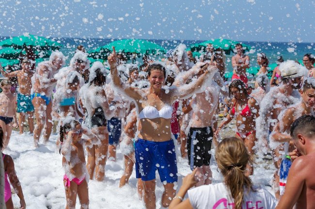Persone partecipano a una festa in spiaggia con schiuma, molte sorridenti e coperte di bolle.