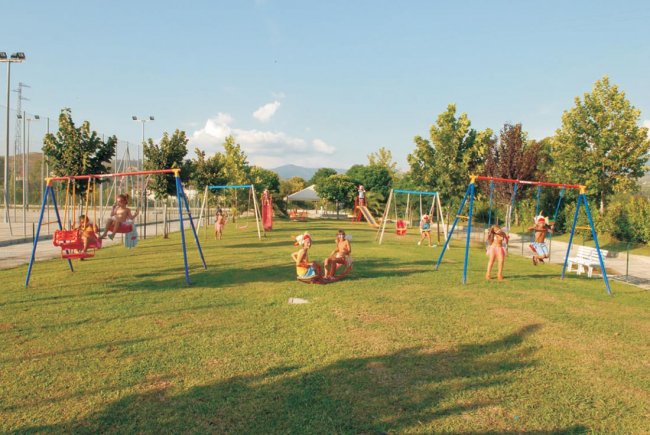 Bambini giocano su altalene in un parco con erba verde e alberi.