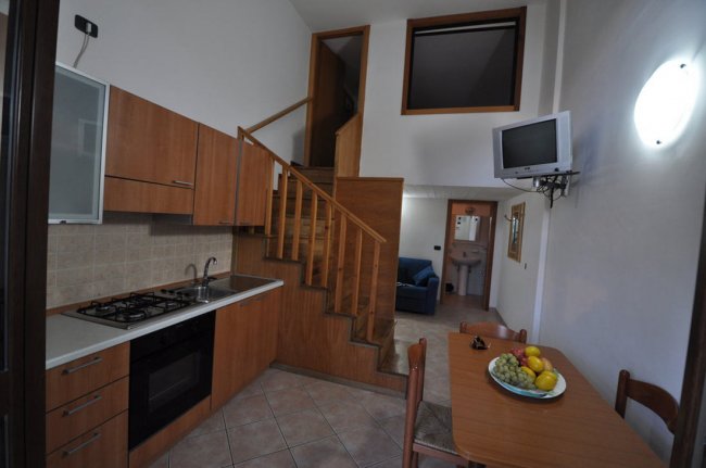 Cucina moderna con mobili in legno, tavolo, scala e TV a muro.