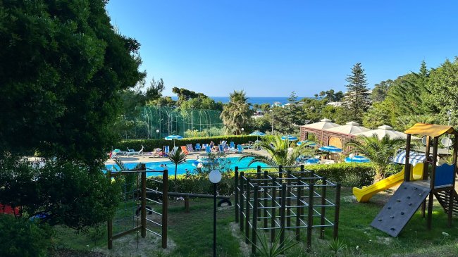 Area giochi con piscina, alberi e vista mare in lontananza.
