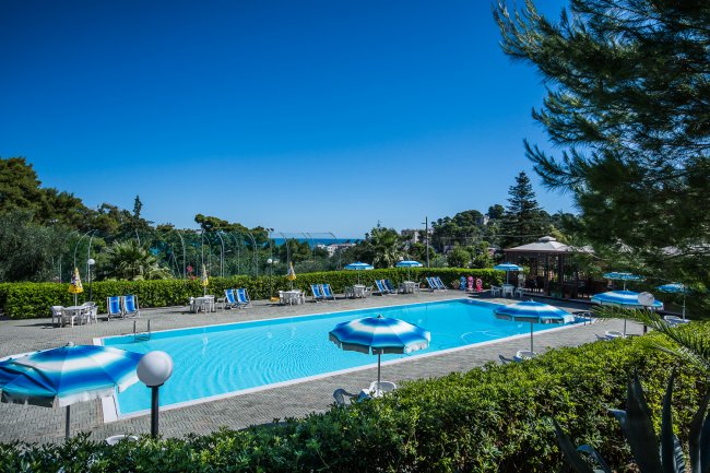 Una piscina all'aperto con ombrelloni blu e bianchi e lettini, circondata da vegetazione.