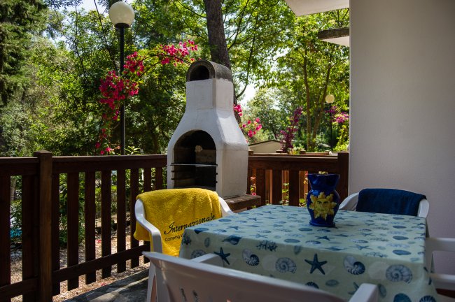 Un patio con tavolo, sedie e barbecue, circondato da fiori rosa e alberi.