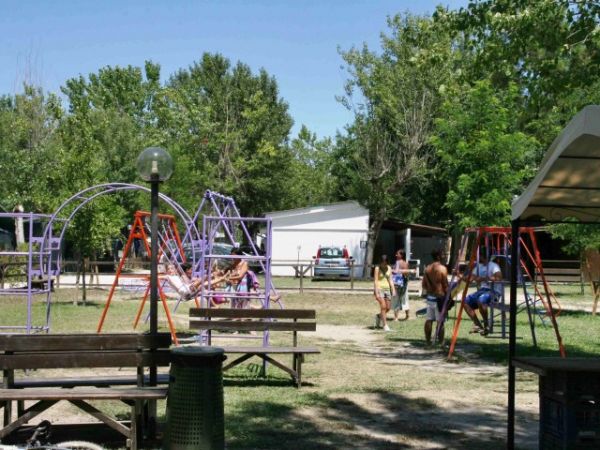 Area giochi con altalene, bambini che giocano, tavoli da picnic e alberi sullo sfondo.